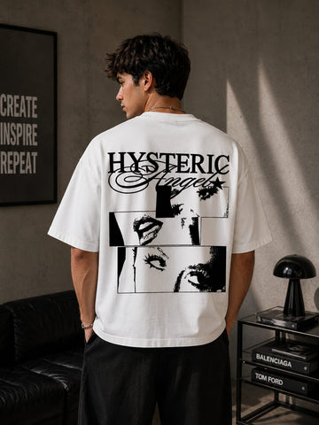 Person wearing a white t-shirt with black graphics and text, standing in a room with a lamp and framed picture.